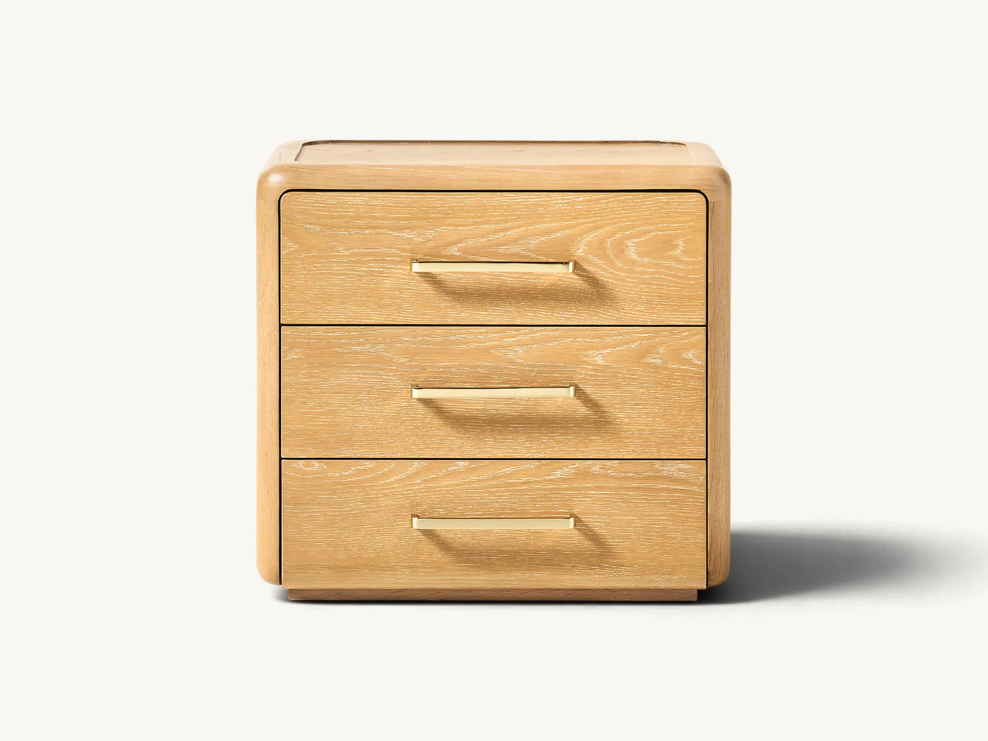 Natural wooden three-drawer nightstand with rounded corners and brass handles set beside a bed