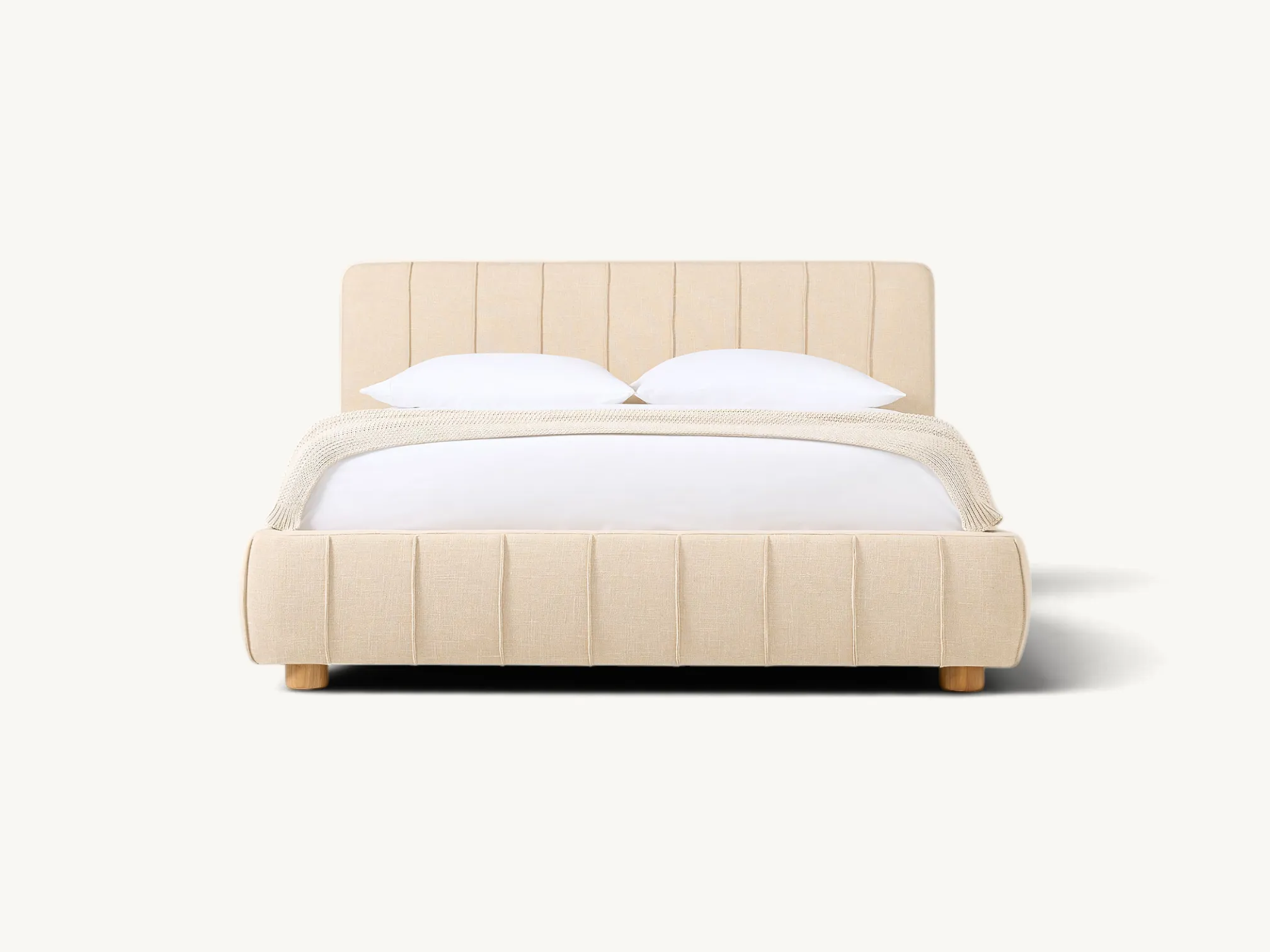 Low-profile upholstered platform bed in warm beige linen fabric, featuring vertical channel pleating detail along the headboard and frame, supported by short cylindrical natural wood legs.